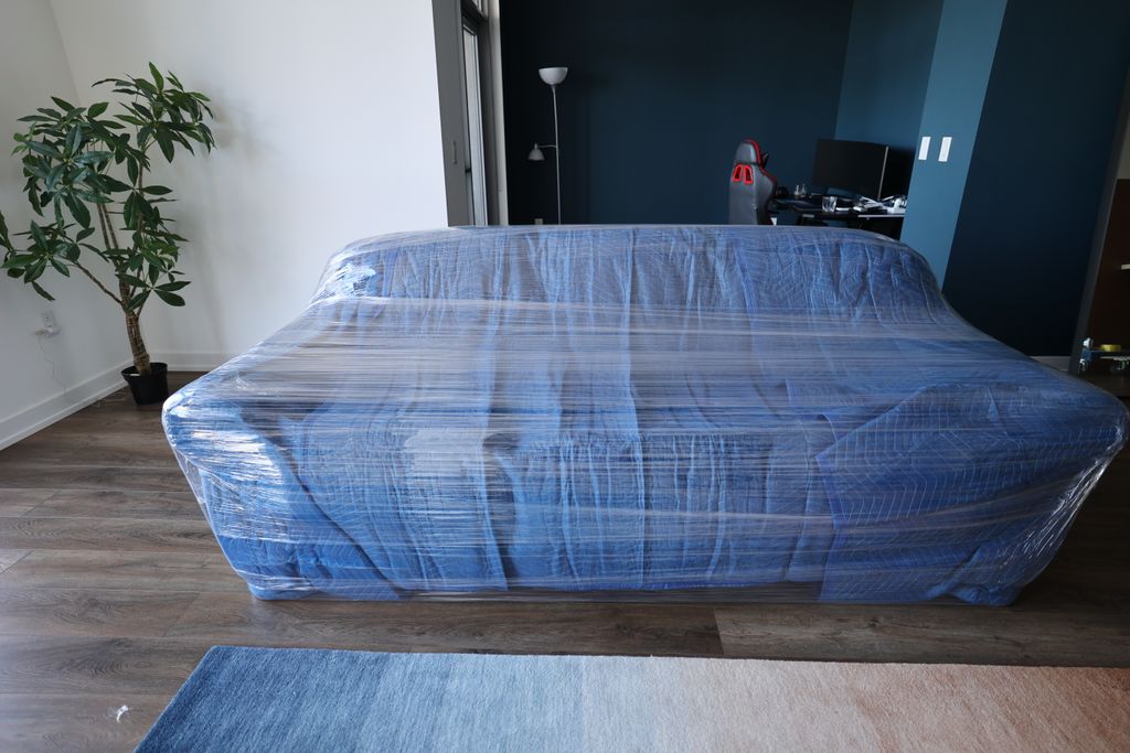 A sofa fully wrapped in a blue padded moving blanket and sealed with clear stretch wrap, standing in full frame in a bright, clean living room, ready to move