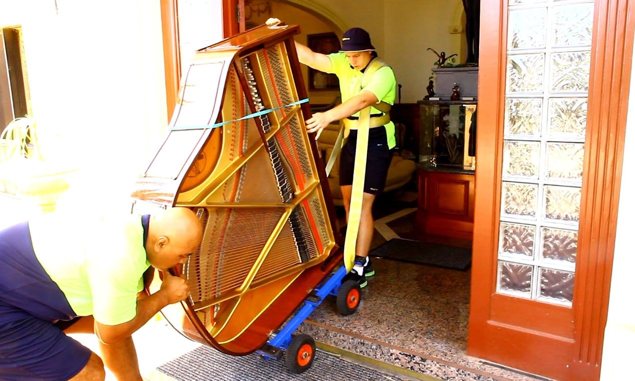 Two movers guiding a grand piano on a piano dolly through a doorway — a real piano relocation in progress