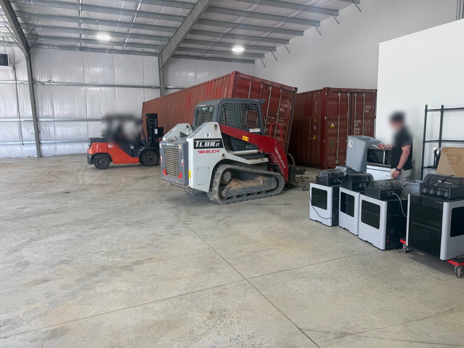 The Flat Rate Movers warehouse — red shipping containers with a Toyota forklift, a Takeuchi track loader and crew working alongside equipment lined up on the warehouse floor