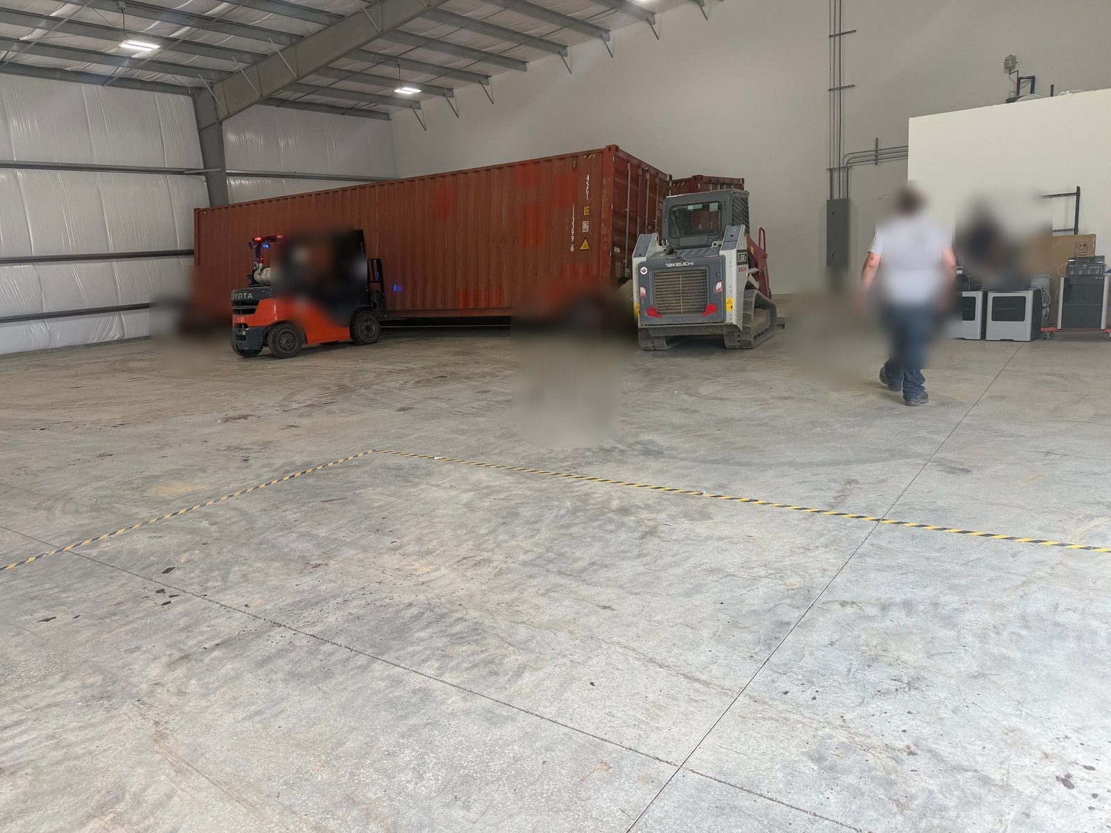 The Flat Rate Movers warehouse and transload operation — crew positioning a red shipping container inside the warehouse with a Toyota forklift and a Takeuchi track loader, with a taped floor zone in the foreground
