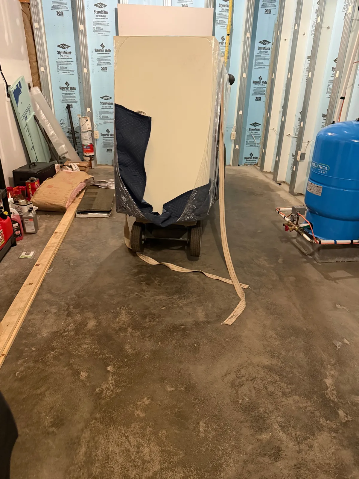 A blanket-wrapped gun safe on a dolly in an unfinished basement corridor, moving straps trailing, a blue water pressure tank to the right