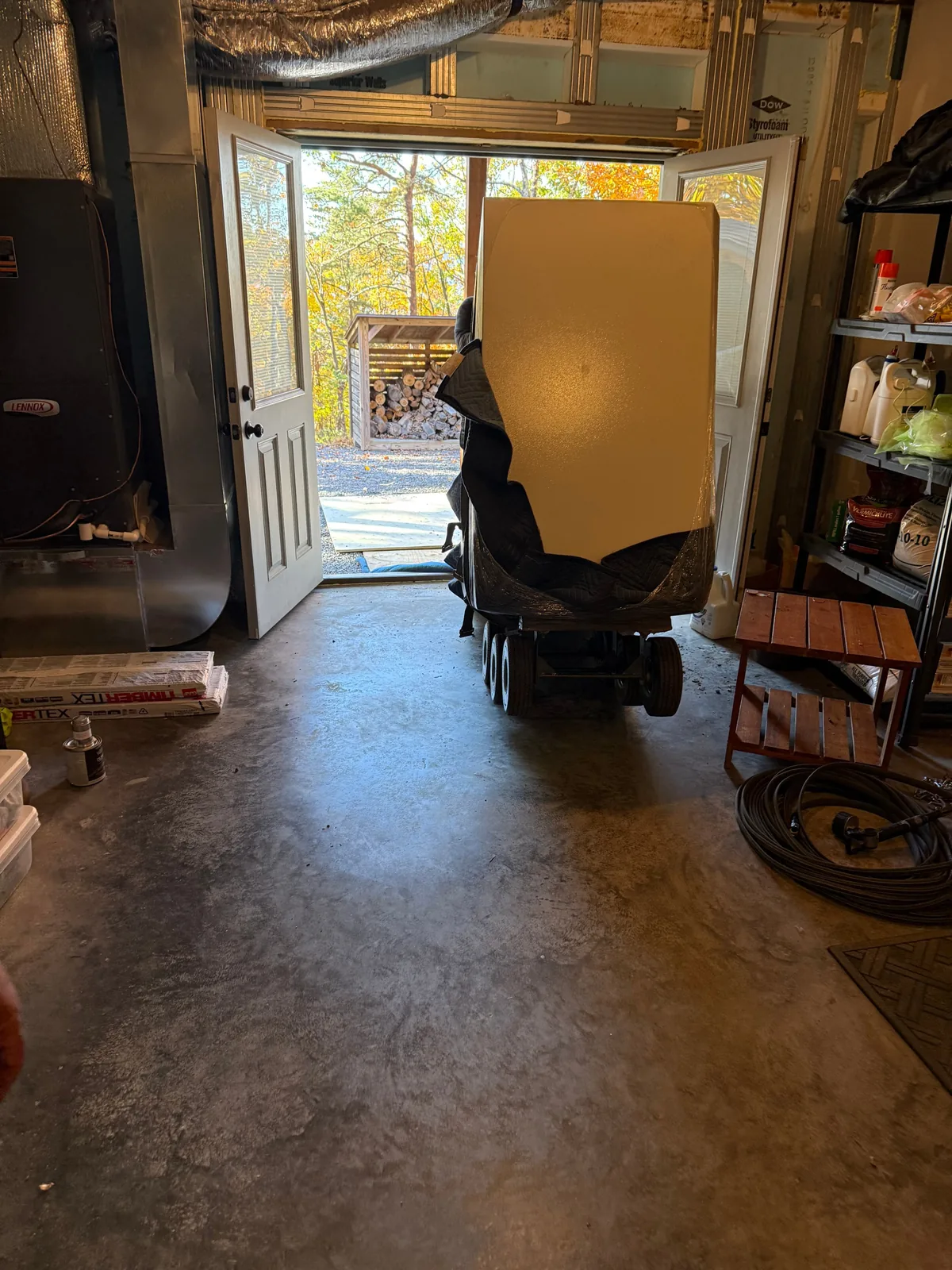 A blanket-wrapped gun safe on an all-terrain dolly at a pair of open double doors, gravel drive and trees visible outside