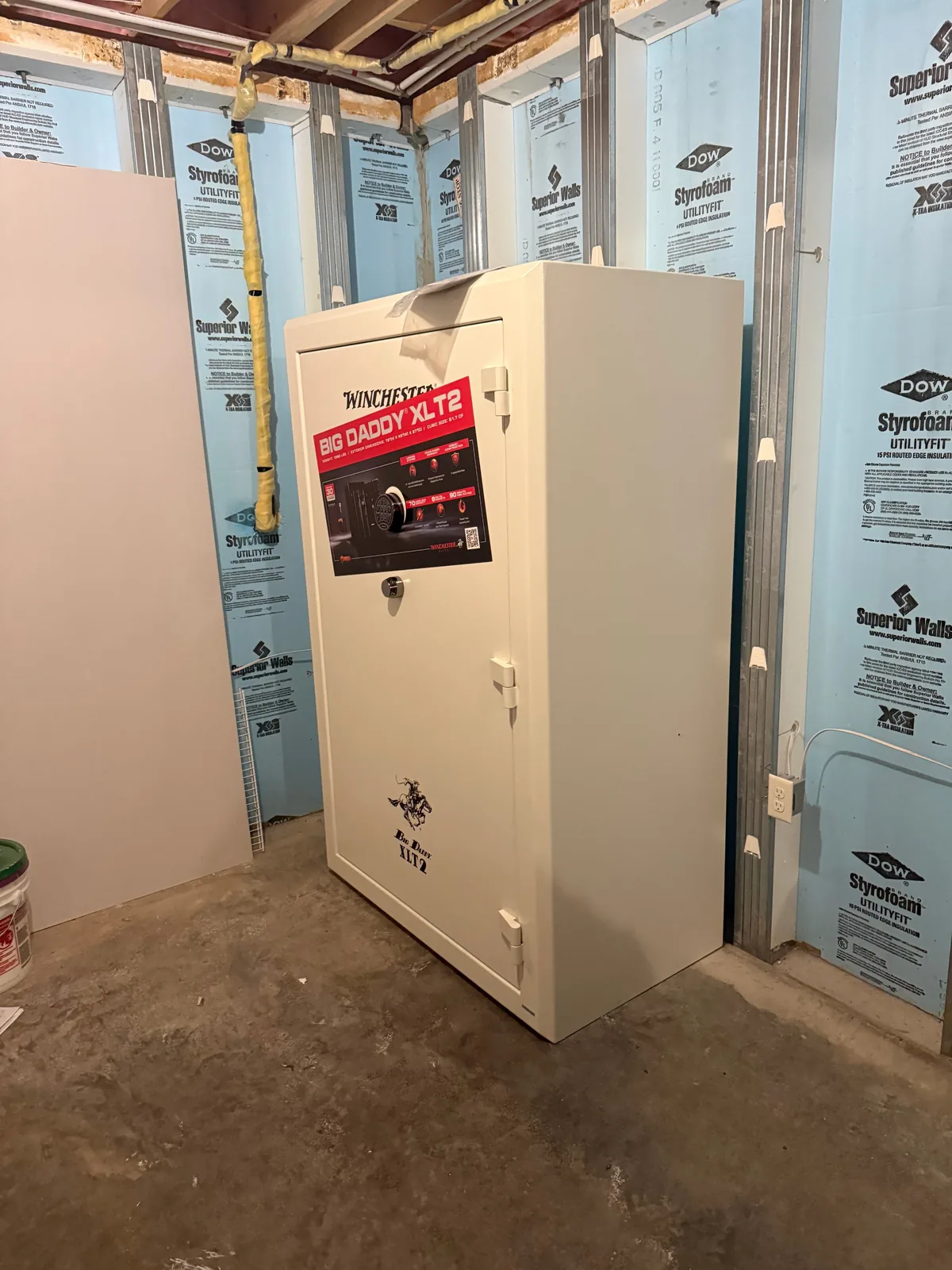 A cream-colored Winchester Big Daddy XLT2 gun safe standing installed in an unfinished basement, manufacturer label on the door