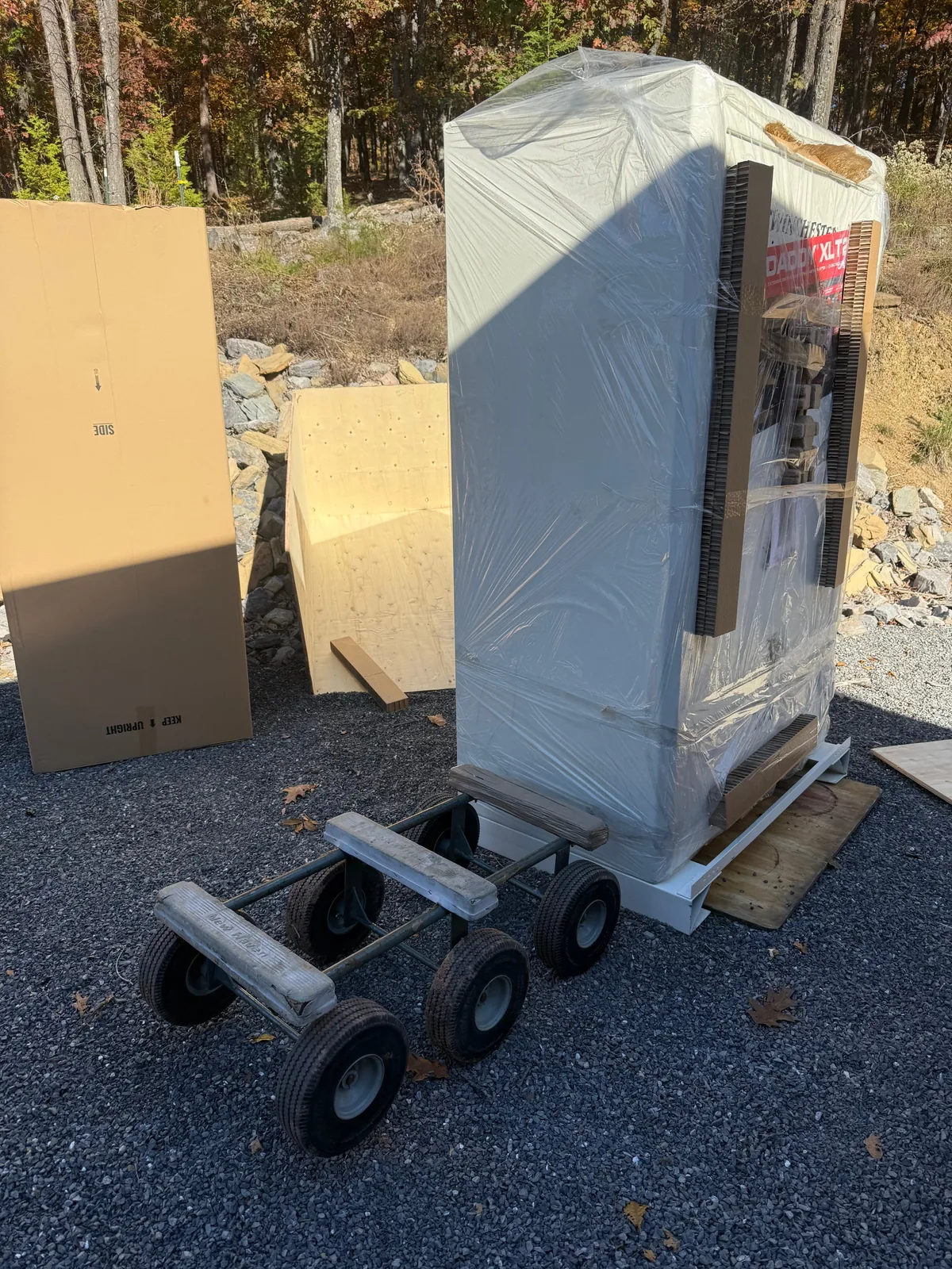 A shrink-wrapped gun safe standing on its shipping skid on a gravel drive, a six-wheel all-terrain dolly in front of it and an opened wooden shipping crate behind it